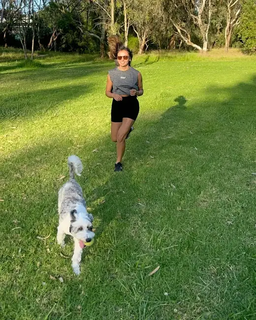 Yen running across a sunny park with a small dog carrying a yellow ball