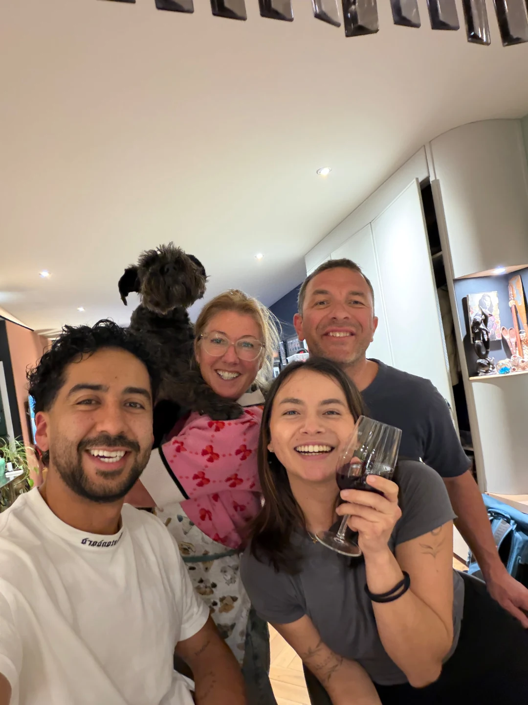 A group of four people posing closely together for a photo inside a home, one holding up a small dark shaggy dog and another raising a glass of red wine