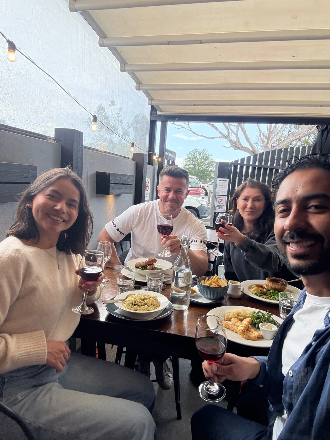 Four people seated around a wooden table on a covered pub patio, raising glasses of red wine in a toast, plates of food between them and string lights overhead