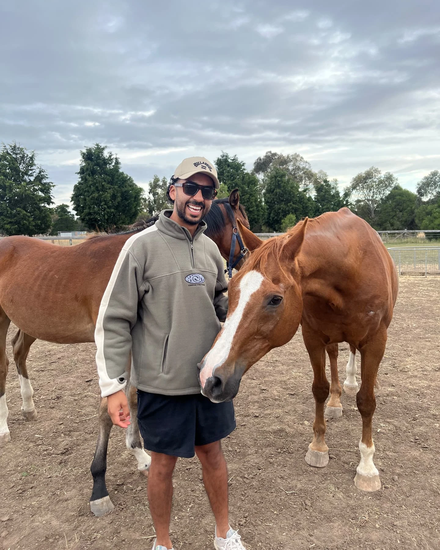 Nico smiling in a dirt riding paddock in a sage green quarter-zip fleece and a cap, a chestnut horse with a wide white blaze leaning its head down beside his waist