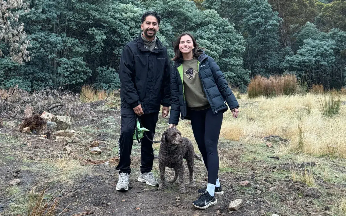 Yen and Nico standing on a dirt track in a forested mountain valley with a dog on a lead between them