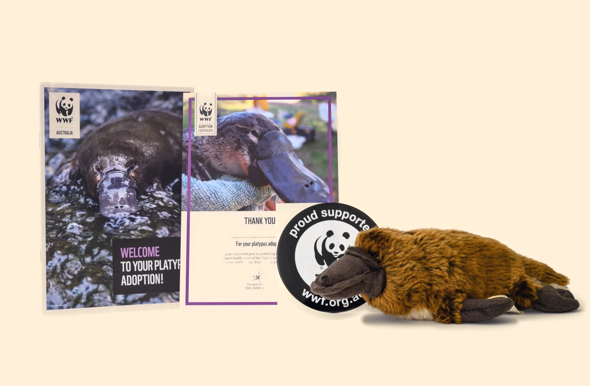 WWF-Australia platypus adoption kit on a cream background: a welcome booklet showing a platypus swimming, an adoption certificate with a close-up photo of a platypus, a round proud supporter sticker and a small brown platypus plush toy.