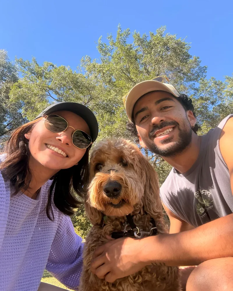 Yen and Nico in caps and sunglasses with a cavoodle in a sunny park