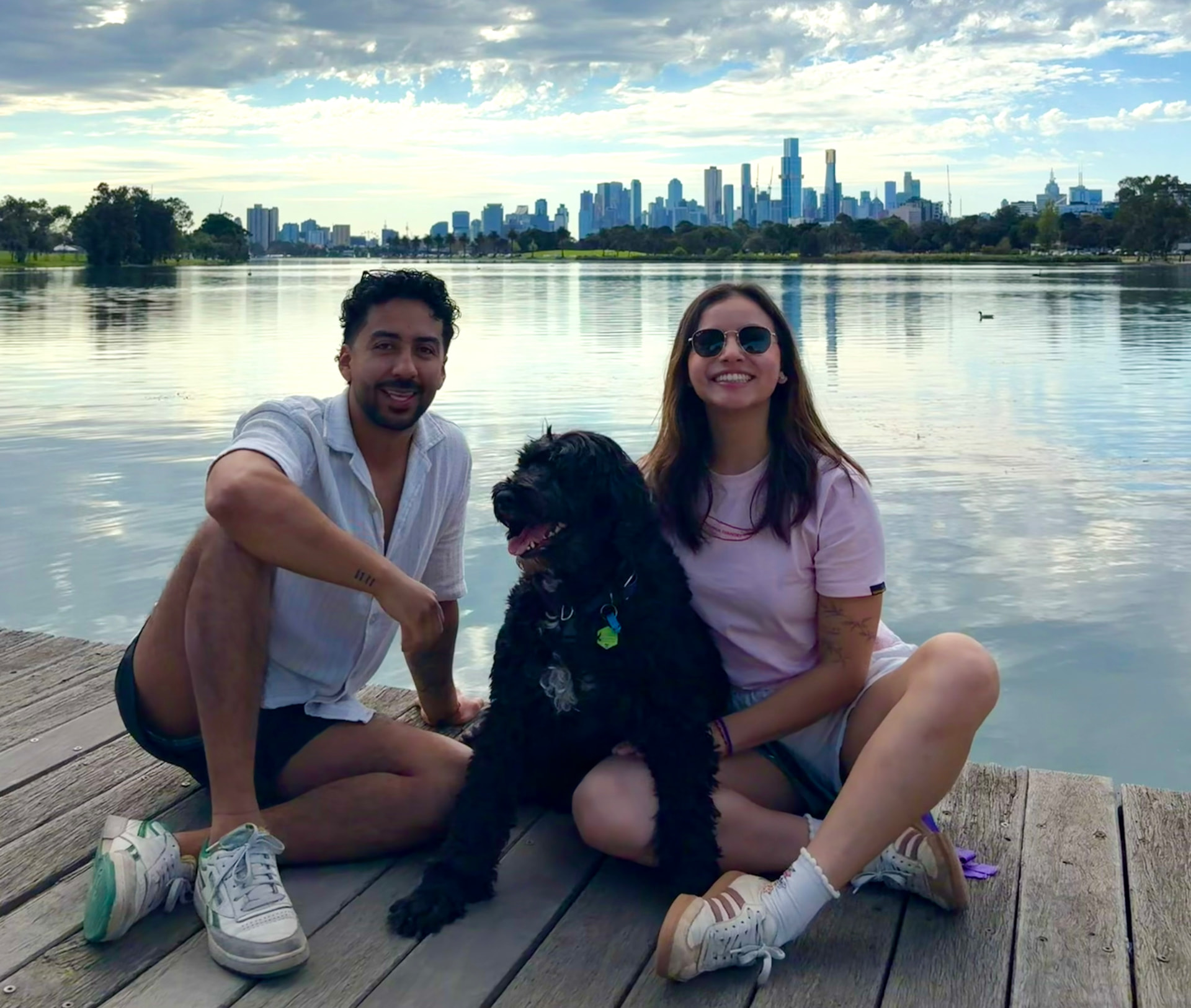 A man and a woman sitting on weathered wooden dock planks with a large shaggy black dog between them, a calm lake and city skyline behind