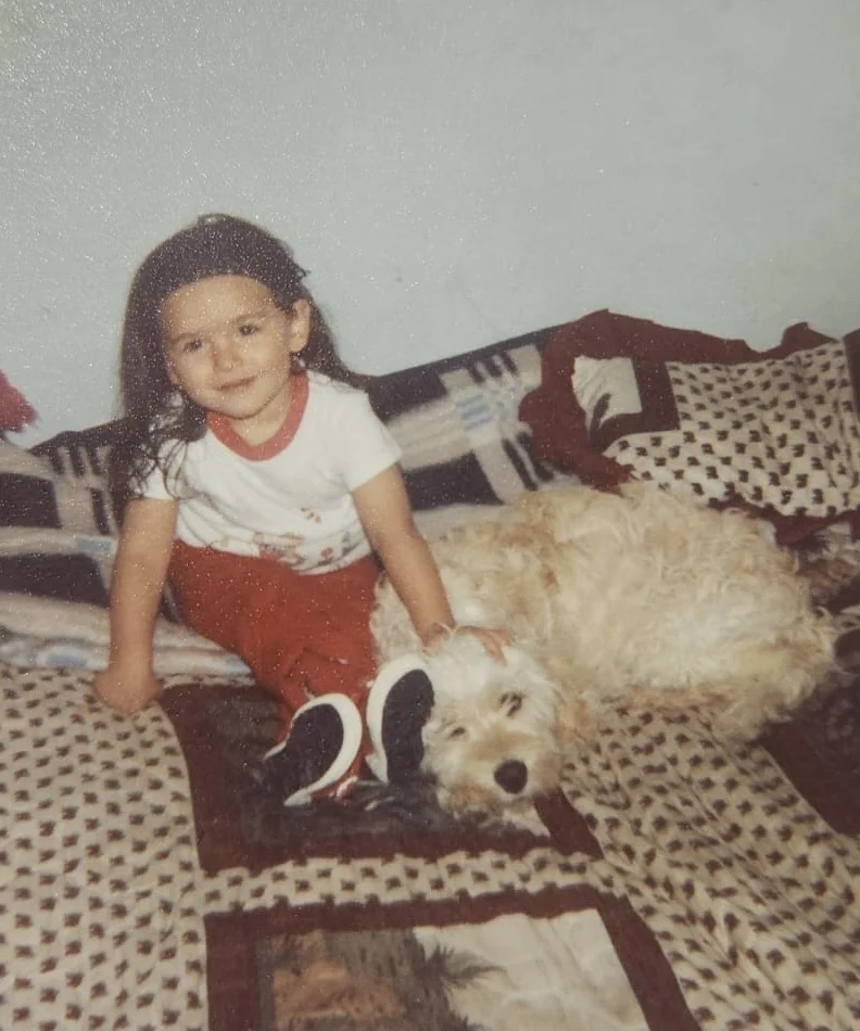 A childhood photo of Yen as a young girl with dark hair, sitting on a bed with a fluffy cream dog lying beside her and her hand resting on the dog's head
