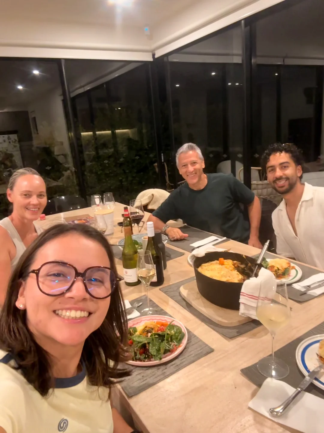 Four people gathered around a home dining table at night, a cast-iron dish of baked food with a golden crust in the centre, plates of salad and glasses of wine in front of them